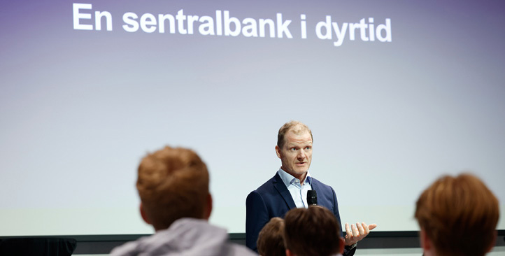 En mann står og prater foran ungdommer med teksten "En sentralbank i dyrtid" på skjerm i bakgrunnen