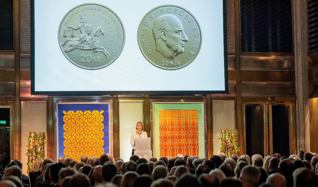 Kvinne på podium foran en forsamling. På storskjerm i bakgrunnen vises for- og bakside av 20-kr minnesmynt