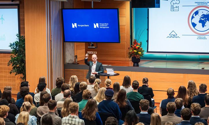 A man leaning on a high round table in front of a stage with two stage monitors in the background, facing an audience.