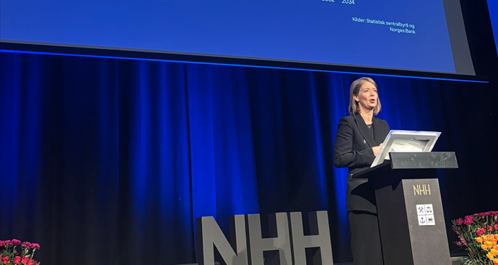 A woman standing at a podium with the letters NHH on the stage and on the podium in front of a stage monitor showing a bar chart.