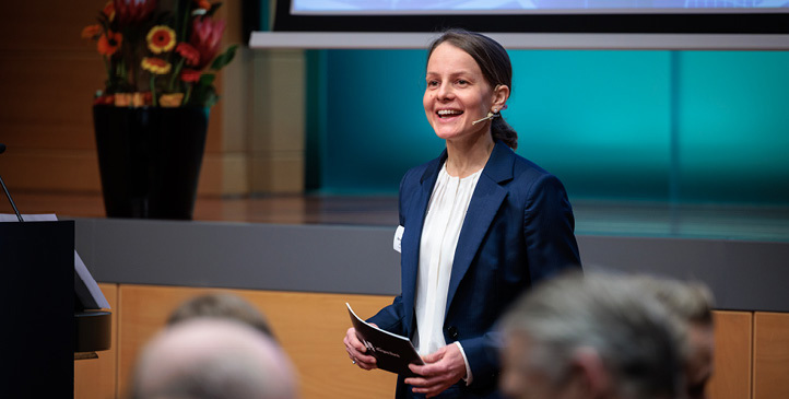 Woman standing, wearing a headset with a microphone, speaking to an audience
