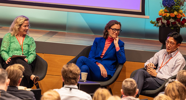 Two women and a man, all sitting in chairs and having a discussion in front of an audience with a stage monitor behind them showing Pride rainbow stripes and the caption "Norges Bank"