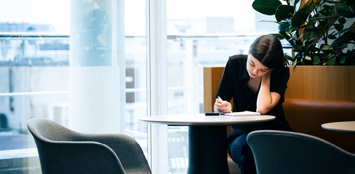 Woman sitting at a small table making notes on paper, next to a potted plant 
