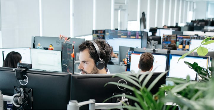 A man wearing headphones looking at two computer screens in an open-plan office with many computer screens and people in the background