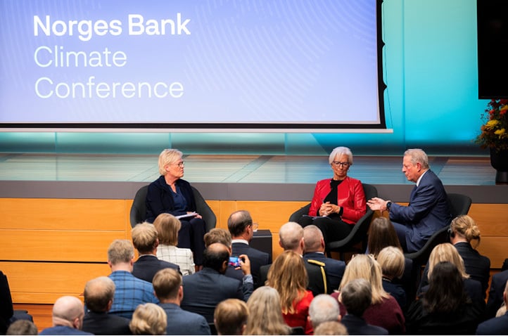 Two women and a man, all sitting in chairs, having a discussion in front of an audience with a stage monitor behind them showing the title Norges Bank Climate Conference