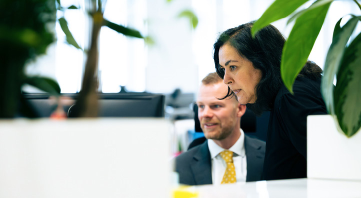 A man and a woman in an open-plan office with green plants looking attentively at a computer screen