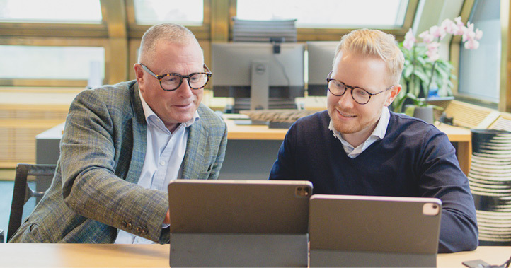 Two men sitting at a table, looking at two laptops