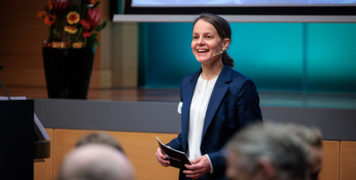 Woman standing, wearing a headset with a microphone, speaking to an audience