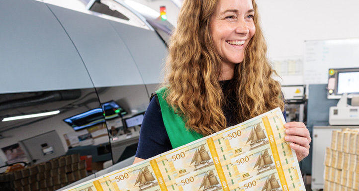 Smiling woman standing by a banknote printing machine and holding a large sheet of 500-krone banknotes