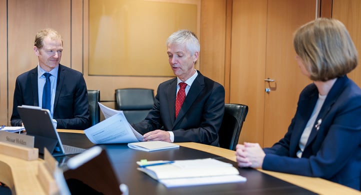 A man and a woman sitting at a meeting table, facing a man between them who is holding some papers 