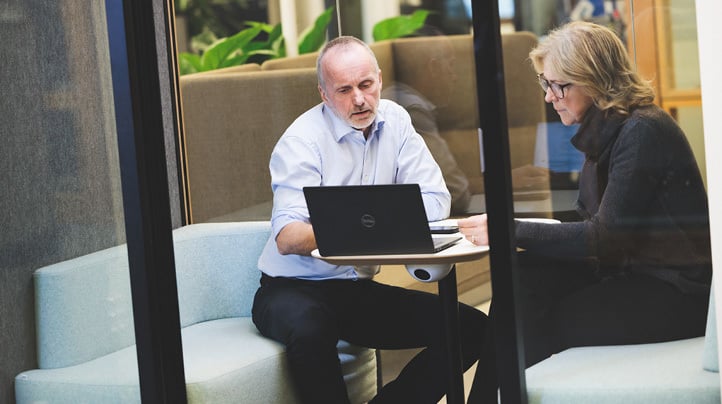 En mann og en kvinne sitter inne i et gjennomsiktig stillerom og ser mot en oppslått bærbar PC på bordet