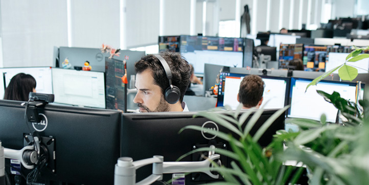 Mann med hodetelefon ser mot to skjermer, og med et kontorlandskap med mange skjermer og personer i bakgrunnen